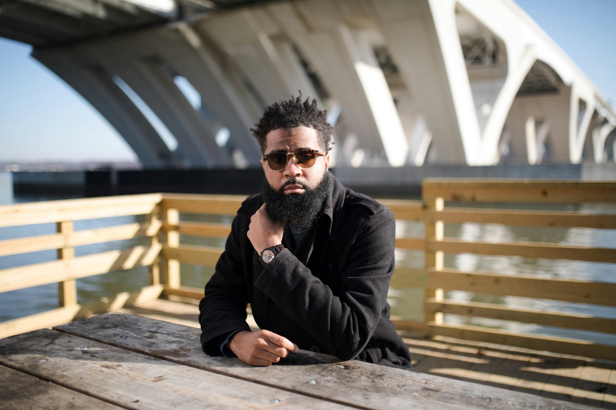 Stylish man sitting on a wooden deck by a bridge, wearing sunglasses and a watch.